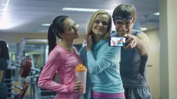 Zwei attraktive, sportliche Mädchen und ein Mann machen Selfie-Bilder mit dem Handy in der Turnhalle. — Stockvideo