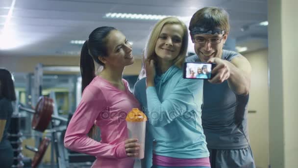 Dos atractivas chicas deportivas en forma y un hombre están haciendo fotos selfie en el teléfono móvil en el gimnasio . — Vídeos de Stock