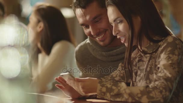 Hombre y mujer se ríen mientras usan un teléfono inteligente y se divierten en un bar . — Vídeos de Stock