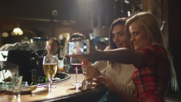 Dos hermosas chicas están haciendo selfies en un teléfono inteligente mientras se divierten en un bar . — Vídeos de Stock