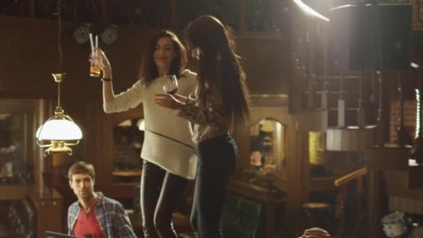 Duas meninas estão dançando com bebidas em uma mesa, enquanto todo mundo está se divertindo juntos em um bar . — Vídeo de Stock