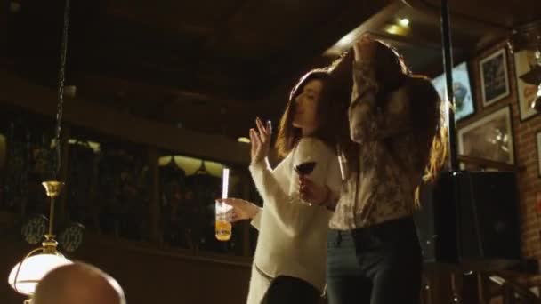 Dwie dziewczyny tańczą z napojami na stole, podczas gdy wszyscy mają dobry czas razem w barze. — Wideo stockowe