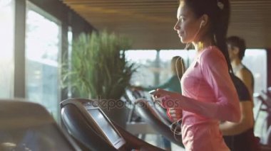 Telefon ve kulaklık ile spor salonunda treadmill çalıştıran çekici beyaz kız.