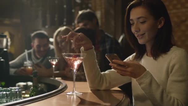 Çekici beyaz kadın bir kokteyl ile bir barda bir akıllı telefon kullanıyor. — Stok video