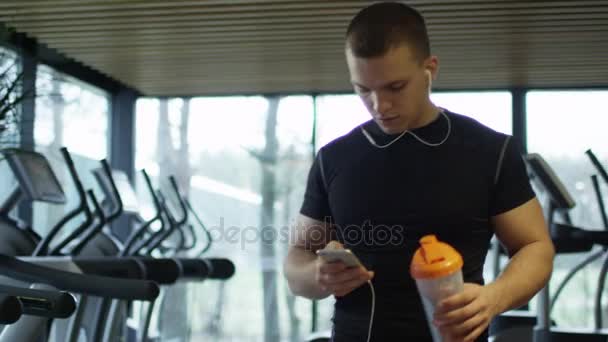 Giovane atletico sta ascoltando musica su uno smartphone mentre cammina in palestra sportiva con tapis roulant . — Video Stock