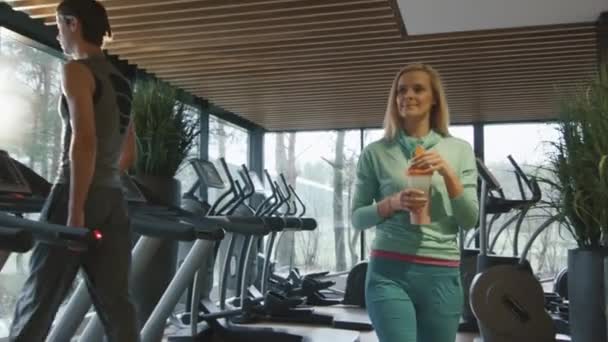 Attraktive blonde kaukasische Mädchen trinkt einen Protein-Shake-Drink, während sie neben einem Laufband in der Sporthalle läuft. — Stockvideo