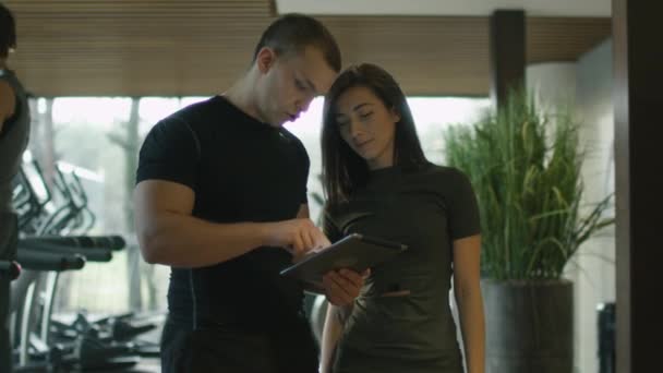 Atletische man en vrouw zijn met behulp van een tablet naast loopbanden in een sportschool sport. — Stockvideo