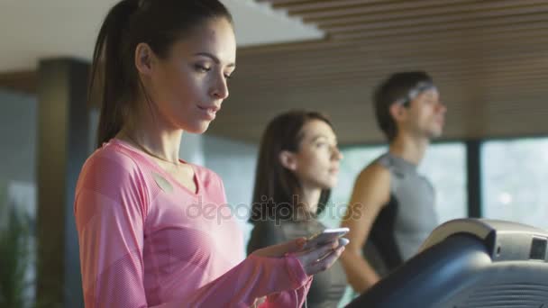 Uygun atletik beyaz kız earphones ve telefon ile spor salonunda koşu bandı üzerinde çalışan. — Stok video