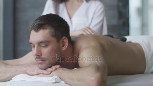 Pohledný muž dostává relaxační masáž kameny od maséra žena ve wellness centru. — Stock video