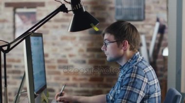 Gözlüklü genç tasarımcı bir çatı katında bir monitör önünde oturan ve bir grafik tablet üzerinde çizim.