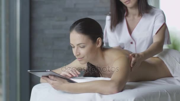 Młoda brunetka kobieta jest przy użyciu tabletu podczas relaksującego masażu w centrum odnowy biologicznej. — Wideo stockowe