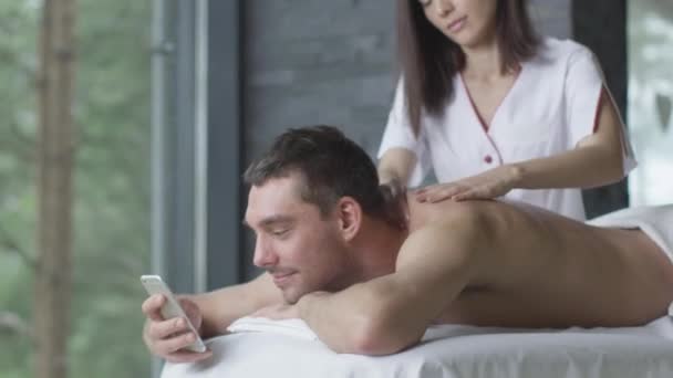 Bonito homem está usando um smartphone durante uma massagem relaxante no centro de bem-estar . — Vídeo de Stock