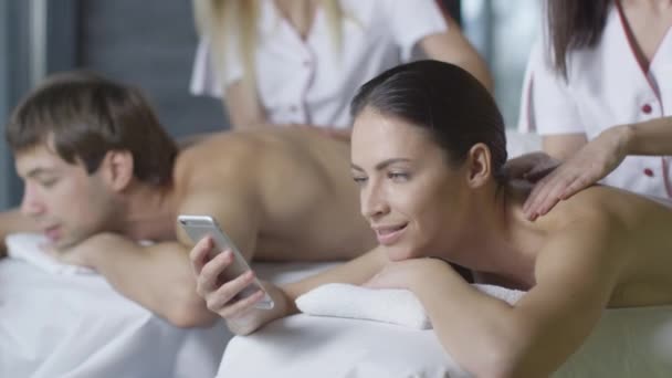 Junge Frau benutzt ein Smartphone während einer entspannenden Paarmassage von Masseurinnen im Wellness-Center. — Stockvideo