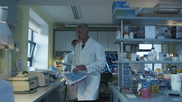 Scienziato di alto livello sta camminando con i documenti in un laboratorio dove i colleghi stanno lavorando . — Video Stock