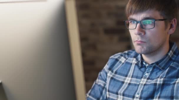 Gros plan d'un jeune homme à lunettes assis devant un moniteur dans un loft et travaillant . — Video