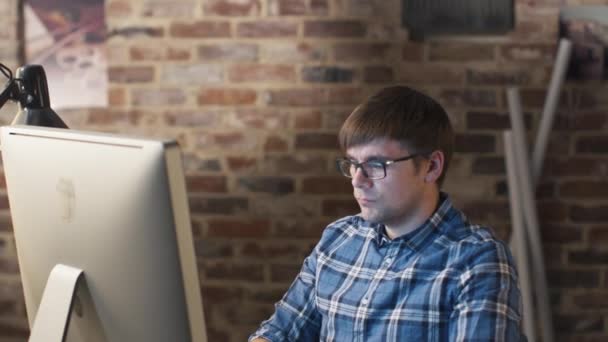 Jovem de óculos está sentado na frente de um monitor em um sótão e trabalhando . — Vídeo de Stock