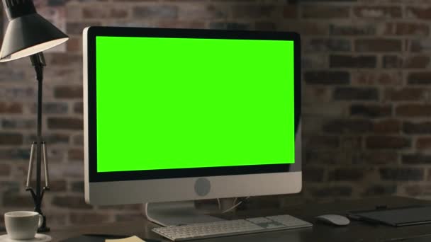 Imágenes de un monitor de computadora con pantalla verde de pie sobre una mesa junto a una tableta, lámpara, taza de café, portátil y ratón en un loft . — Vídeos de Stock