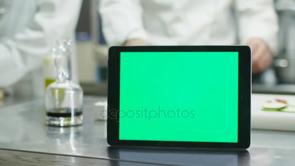 Tablet computador com uma tela verde mock-up está de pé sobre uma mesa em uma cozinha comercial com chefs preparando alimentos em segundo plano . — Vídeo de Stock