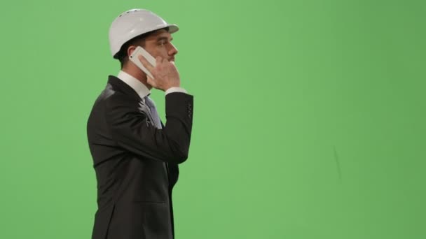 Бизнесмен в каске и в костюме разговаривает по телефону на макете зеленого экрана на заднем плане . — стоковое видео