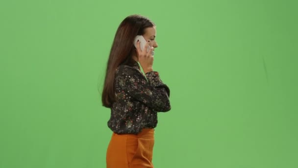 Casual νεαρή μελαχρινή κοπέλα περπάτημα και να μιλάμε στο τηλέφωνο για μια μακέτα πράσινη οθόνη στο φόντο. — Αρχείο Βίντεο