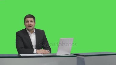 Media yayıncı bir masada oturan ve bir mock-up üzerinde konuşurken arka planda yeşil ekran.