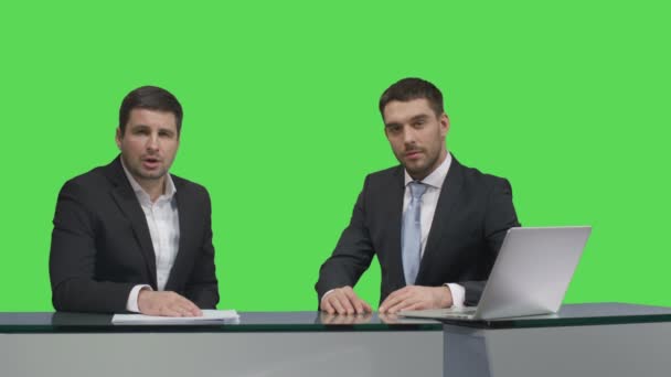 Two media broadcasters are sitting at a table with a laptop and talking on a mock-up green screen in the background. — Stock Video