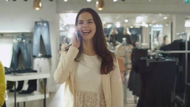 Cheerful young brunette girl is walking though a clothing store while talking on the phone. — Stock Video