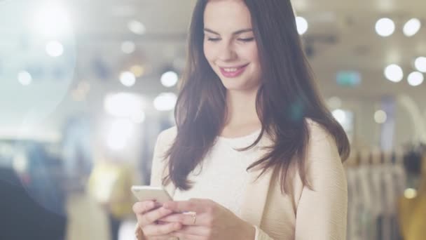 Jovem morena feliz está de pé em uma loja de departamento e mensagens de texto em um smartphone . — Vídeo de Stock