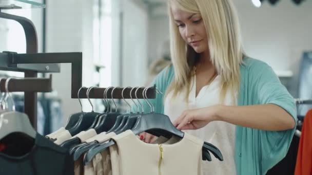 Menina loira está comprando vestidos e procurando um tamanho certo em uma loja de departamento . — Vídeo de Stock