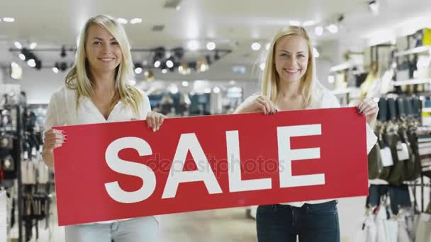 Две счастливые блондинки держат плакат с распродажей в магазине одежды . — стоковое видео