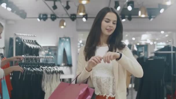 Счастливая юная брюнетка ходит через магазин одежды, проверяя уведомления на своих умных часах . — стоковое видео