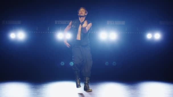 Caucásico bailarín masculino en un chaleco y pantalones anchos está bailando en un escenario oscuro ahumado . — Vídeos de Stock