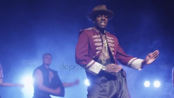 Uomo afroamericano in cappello e costume sta guidando un gruppo di ballerini mentre canta su un palco buio con luci . — Video Stock