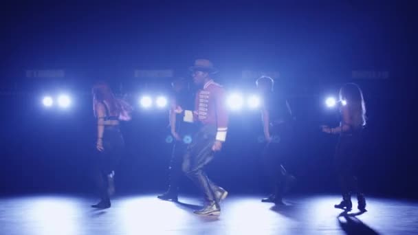 Αφρικανική αμερικανική άνδρα σε ένα καπέλο και κοστούμι οδηγεί μια ομάδα χορευτών, ενώ το τραγούδι σε μια σκοτεινή σκηνή με τα φώτα. — Αρχείο Βίντεο
