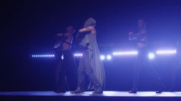 Africano americano macho em um capuz leva um grupo de dançarinos enquanto canta em um palco escuro com luzes . — Vídeo de Stock