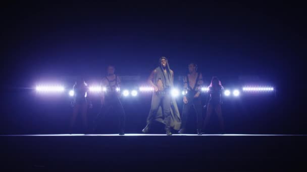 African american male in a hoodie leads a group of dancers while singing on a dark stage with lights. — Stock Video