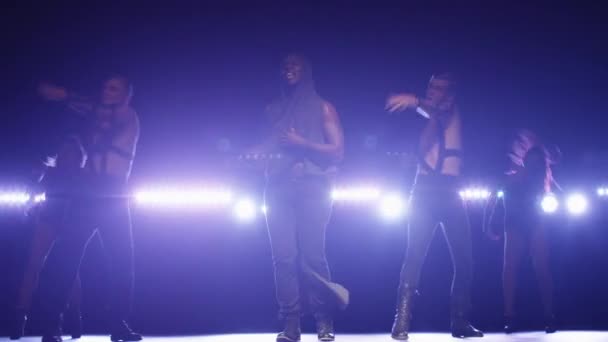 Afrikanischer amerikanischer Mann im Kapuzenpulli führt eine Gruppe von Tänzern an, während er auf einer dunklen Bühne mit Lichtern singt. — Stockvideo