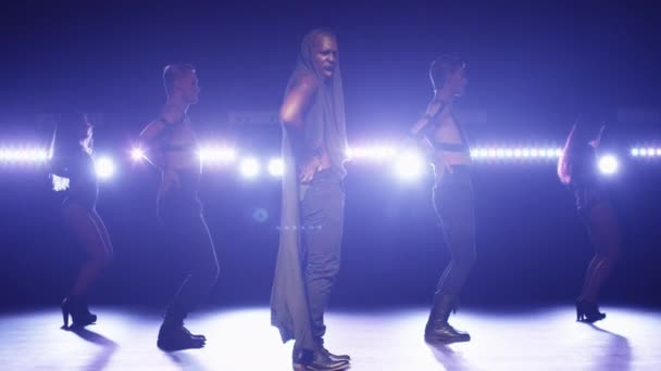 Homme afro-américain en sweat à capuche dirige un groupe de danseurs tout en chantant sur une scène sombre avec des lumières . — Video
