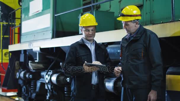 Dos técnicos con sombreros duros conversan al lado de Train. Un técnico que sostiene la tableta PC . — Vídeo de stock