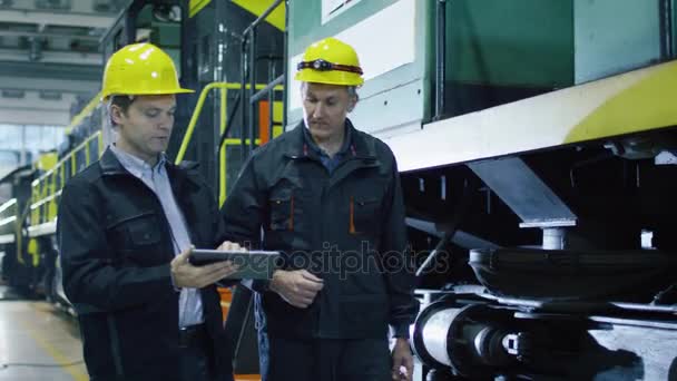 Twee technici in harde hoeden hebben gesprek naast trein. Een technicus bedrijf Tablet Pc. — Stockvideo