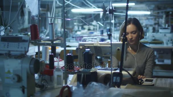 Trabajadora de prendas de vestir joven está cosiendo en una fábrica de ropa mientras escucha música en su teléfono inteligente durante un turno de noche . — Vídeo de stock