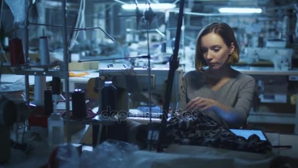 Mujer joven trabajadora de la confección está mirando una tableta y cosiendo en una fábrica de ropa durante un turno de noche . — Vídeos de Stock
