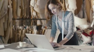 Kadın giyim Tasarımcısı bir stüdyo masanın üzerine eğilerek bir dizüstü bilgisayarda çalışıyor.