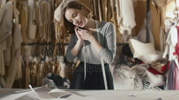 Jonge vrouw kleding ontwerper is praten over een telefoon naast een tabel studio. — Stockvideo