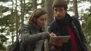 Uzun saçlı erkek ve karanlık sonbahar giysileri esmer kadın bir ormanda ayakta ve tablet kullanma.