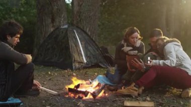 Bir grup insan içecekler ile bir kamp ateşi yanında oturmak ve tablet bir ormanda kullanılması.