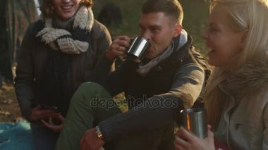 Arkadaş grubu termos sıcak içecekler ile bir kamp ateşi yanında bir ormanda oturup konuşmak.