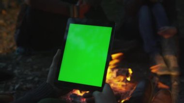 Kişi bir tablet ile bir kamp ateşi yanında yeşil ekran mock-up portre modunda akşamları tutuyor.