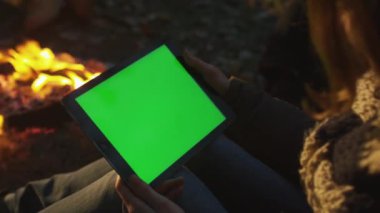 Kız bir tablet ile bir kamp ateşi yanında yeşil ekran mock-up manzara modunda akşamları tutuyor.