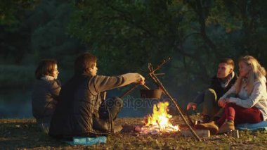 Bir grup insan bir güneşli gün ve aşçı bir kamp ateşi yanında oturuyor.